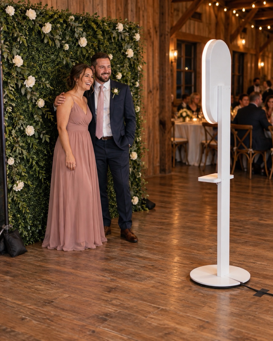 A couple posing in front of a greenery wall backdrop while the white photo booth faces them at a barn wedding