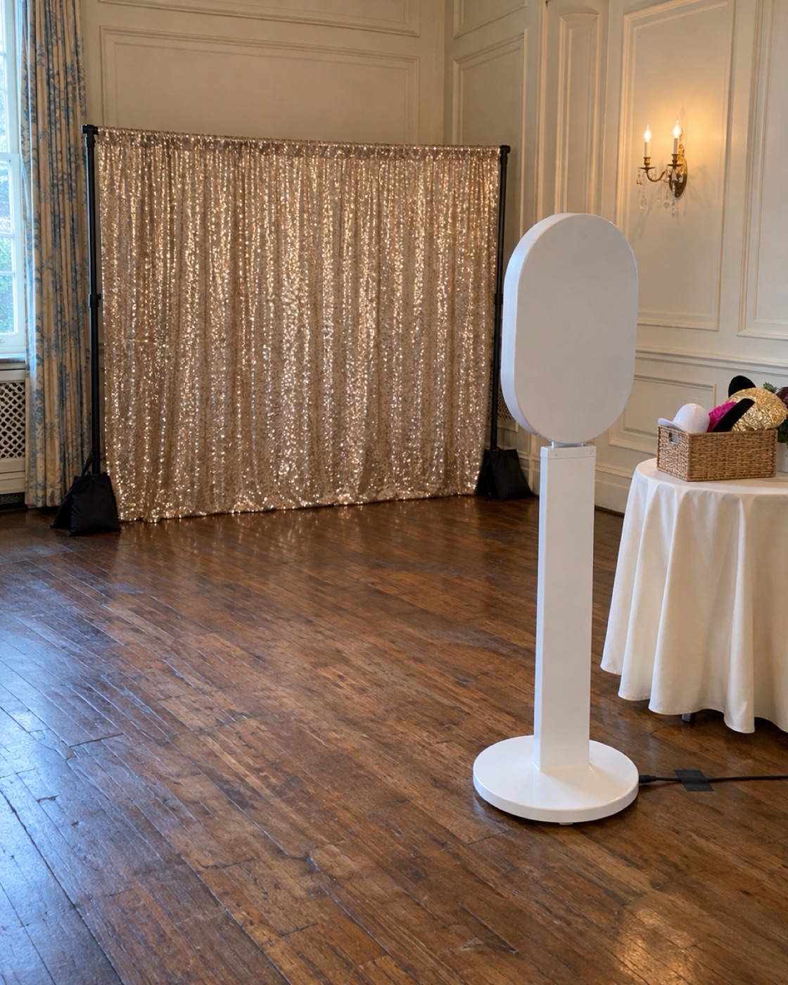 The white photo booth, seen from behind, aimed at a champagne sequin backdrop in the corner of a ballroom, with a basket of props beside it