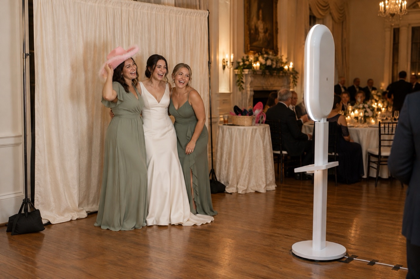 A bride and two bridesmaids posing in front of an ivory drape backdrop while the white photo booth faces them